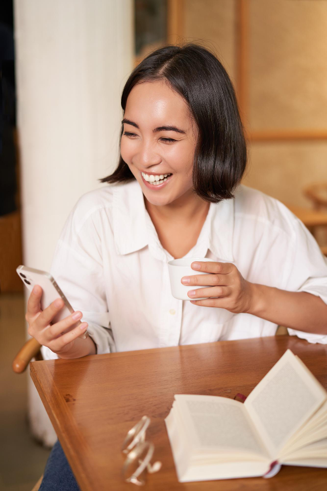 Happy asian woman sits in cafe with cup of coffee and book answer video call on smartphone laughing Related tags: