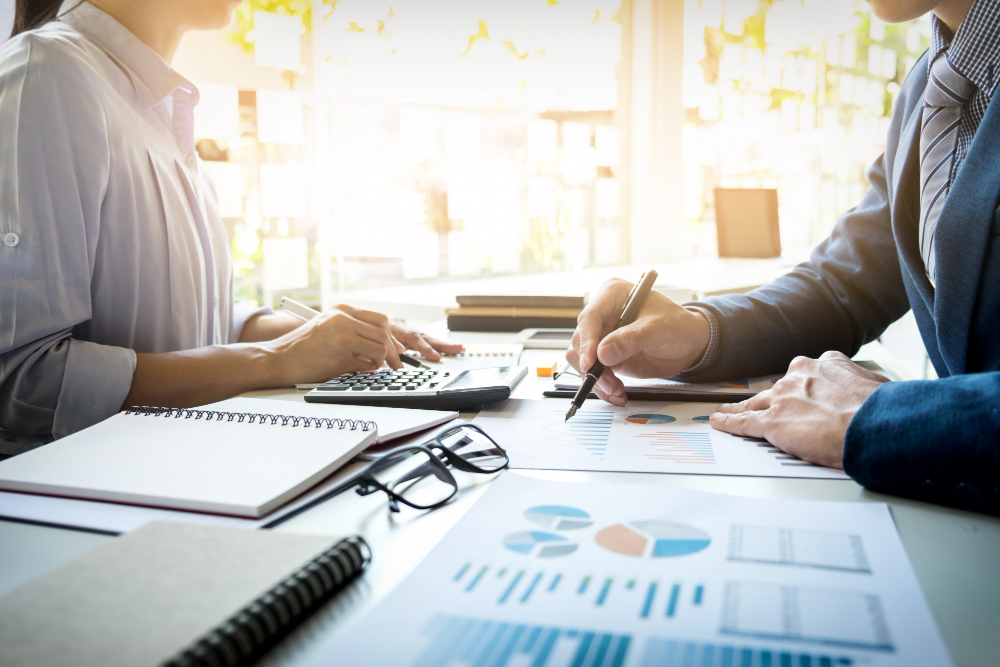 Two business professionals analyzing financial data and graphs on a desk with a calculator, pen, notebook, and glasses.