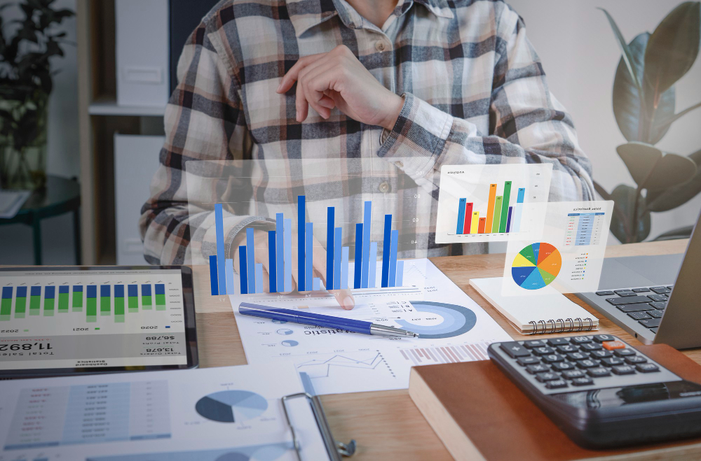 Person in plaid shirt analyzing data on transparent screen with various bar graphs, pie charts, and a laptop on wooden desk.