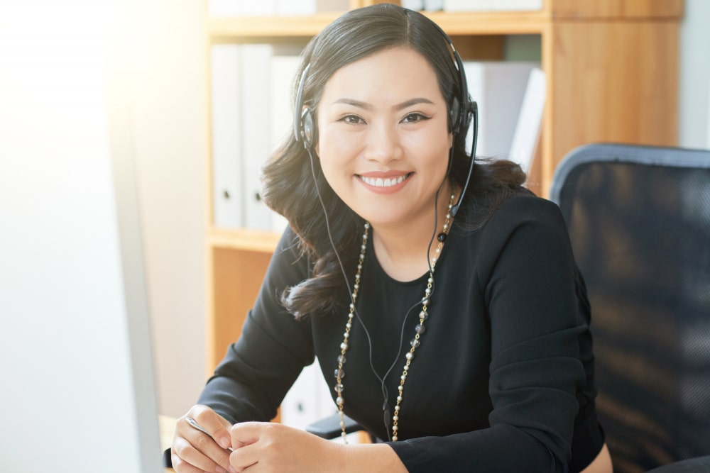 A professional woman wearing a headset, sitting at a desk with a blurred logo of "IO Outsourcing Services" visible in the foreground. She has long dark hair and is wearing a black top with a string of beads. A bright office environment with cabinets and papers can be seen in the background.
