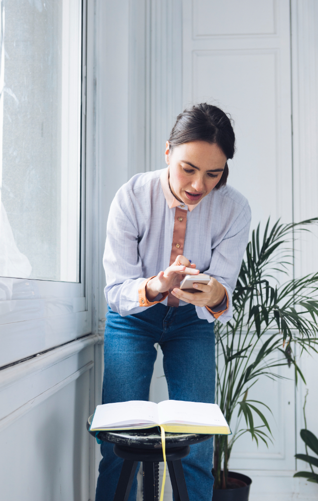 A person in a light blue shirt and dark blue jeans standing by a window, looking at a smartphone with a notebook on a small black stool in front of them, in a room with a potted plant.